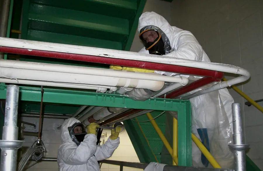 Two workers in protective suits and respirators removing asbestos lagging from pipework
